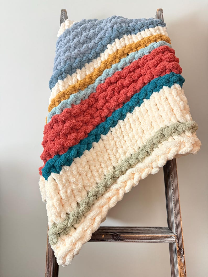 May include: A chunky knit throw blanket featuring diagonal stripes in cream, dusty blue, mustard yellow, terracotta red, teal, and sage green. The blanket is crafted from thick, soft yarn with a prominent, textured loop stitch pattern throughout.