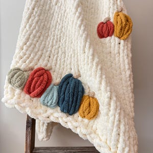 May include: A chunky knit cream-coloured blanket featuring several small, plush, three-dimensional pumpkins in various colours including sage green, burnt orange, light blue, dark blue, mustard yellow, and rust red attached to the surface.