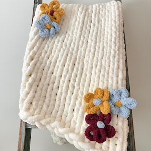 May include: A chunky knit cream-coloured blanket featuring thick, soft yarn loops. The blanket is decorated with five crocheted flower appliques in mustard yellow, light blue, and deep burgundy, each with a contrasting centre colour.