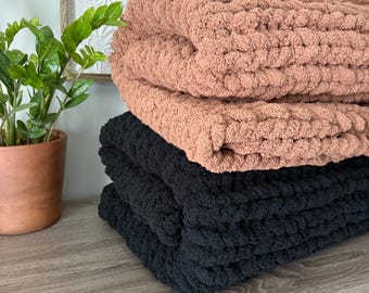 May include: A stack of two chunky knit blankets, one in a muted terracotta color and one in solid black. The blankets feature a thick, soft, looped yarn texture, folded neatly to display the dense, hand-knitted construction of the fabric.