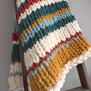 May include: A chunky knit blanket with a chevron design, displayed on a wooden ladder. The blanket has stripes of cream, red, teal, blue, and mustard yellow. The edges are finished with a thick, cream border.