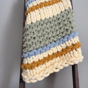 May include: A chunky knit blanket in cream, sage green, blue, and mustard yellow stripes is draped over a wooden ladder. The soft, textured blanket is a cosy home decor item, perfect for adding warmth and style to any space.