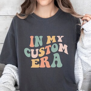 May include: A dark gray t-shirt with the text "IN MY CUSTOM ERA" in a retro font with colorful letters. The letters are in shades of green, pink, orange, and yellow.