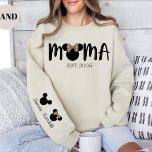 May include: Sand-colored sweatshirt with the word "MOMA" in black, featuring a Mickey Mouse head with a leopard print bow. The text "EST.2005" is below. The left sleeve has Mickey Mouse heads and the names "James Scarlett".