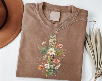 May include: A brown t-shirt with a floral cross design. The cross is composed of various flowers in shades of white, pink, yellow, and green. A silver necklace with a small pendant rests on the shirt. A brown hat is visible in the upper left corner.