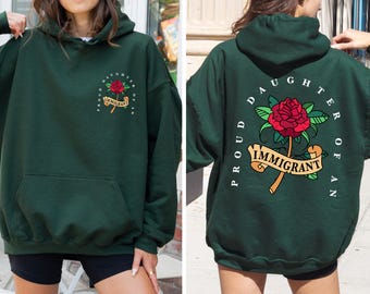 May include: A dark green hooded sweatshirt with the text "Proud Daughter of an Immigrant" and a red rose with green leaves and a banner that says "Immigrant".