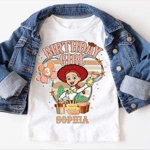 May include: White t-shirt featuring a cartoon image of a character with red hair, a white shirt, and a green skirt. The shirt has the text "BIRTHDAY GIRL" and the name "SOPHIA" printed on it. The shirt is paired with a blue denim jacket.