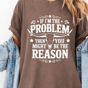 Peut inclure: T-shirt marron avec l'inscription blanche « IF I'M THE PROBLEM THEN YOU MIGHT BE THE REASON ». Le motif comprend un crâne de taureau et des étoiles. Le t-shirt est de style décontracté.