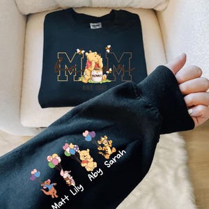 A person's hand is holding a black sweatshirt with a design featuring Winnie the Pooh and the names of three individuals, Matt, Abby, and Lily.