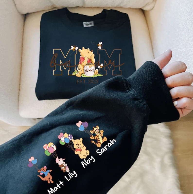 A person's hand is holding a black sweatshirt with a design featuring Winnie the Pooh and the names of three individuals, Matt, Abby, and Lily.