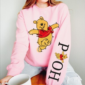 A woman wearing a pink sweatshirt with a cartoon character printed on it, sitting on a wooden stool and holding a white mug.