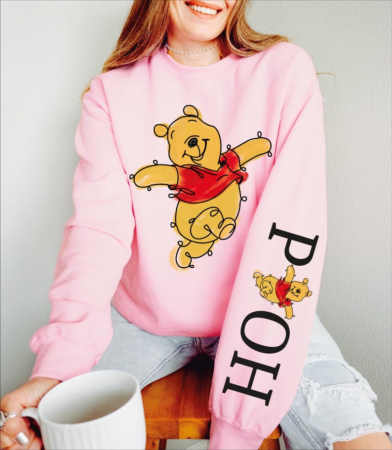 A woman wearing a pink sweatshirt with a cartoon character printed on it, sitting on a wooden stool and holding a white mug.