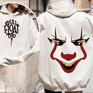 May include: Cream-colored hoodie with a front pocket and hood. The front features the text "YOU'LL FLOAT TOO" in a black, stylized font. The back displays a graphic of a clown face with red and brown accents.