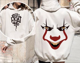 May include: Cream-colored hoodie with a front pocket and hood. The front features the text "YOU'LL FLOAT TOO" in a black, stylized font. The back displays a graphic of a clown face with red and brown accents.