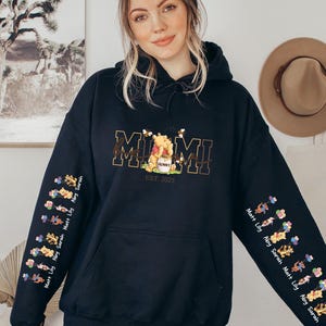 A woman wearing a black hoodie with a graphic design on the front, standing in front of a wall with a framed picture and a hat.