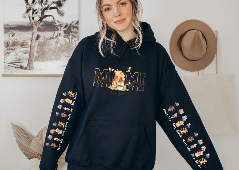 A woman wearing a black hoodie with a graphic design on the front, standing in front of a wall with a framed picture and a hat.
