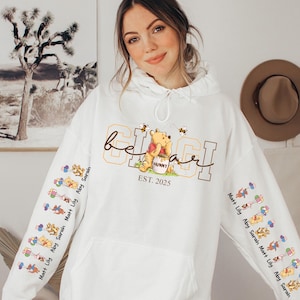 A woman wearing a white hoodie with a Winnie the Pooh design and text on it, posing in front of a wall with a painting and a hat.