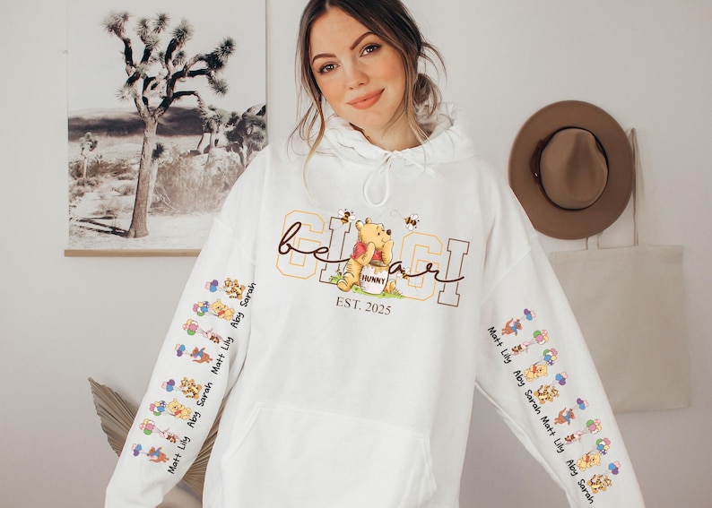 A woman wearing a white hoodie with a Winnie the Pooh design and text on it, posing in front of a wall with a painting and a hat.