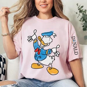 A woman wearing a pink t-shirt with a cartoon image of Donald Duck is sitting on a couch.