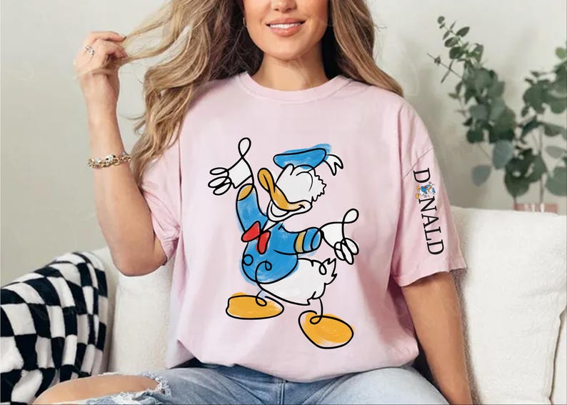 A woman wearing a pink t-shirt with a cartoon image of Donald Duck is sitting on a couch.