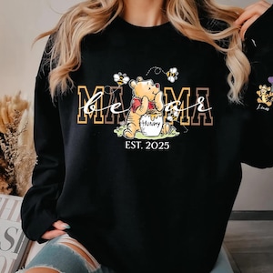 A woman wearing a black sweatshirt with a Winnie the Pooh-themed design, including the character's name and the year "EST. 2025".