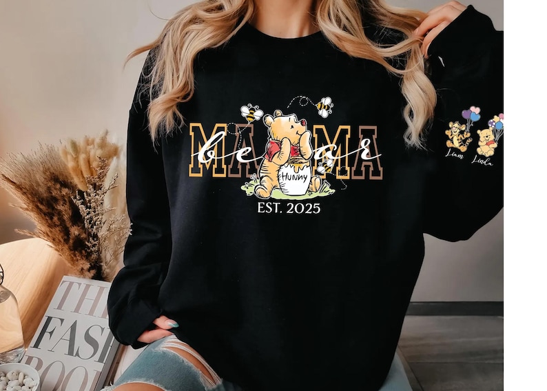A woman wearing a black sweatshirt with a Winnie the Pooh-themed design, including the character's name and the year "EST. 2025".