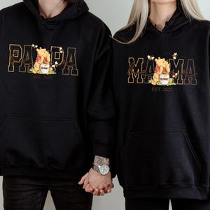 Two people wearing black hooded sweatshirts with the text "PAPA" and "MAMA" printed on them.