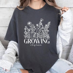 May include: A dark grey t-shirt with a white floral design and the text "GROWING a tiny human".