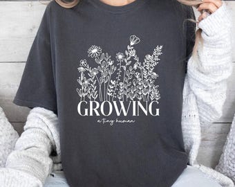 May include: A dark gray t-shirt with a white floral design and the text "GROWING a tiny human".