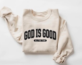 May include: A cream-colored sweatshirt with the text "GOD IS GOOD" in a black, collegiate-style font. Below the main text is the phrase "ALL THE TIME" in a smaller, black font. The sweatshirt is folded, with the sleeves visible.