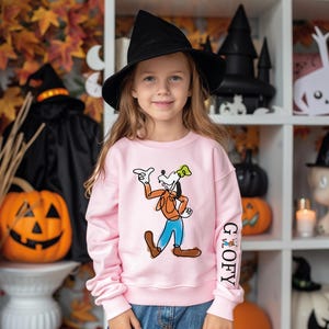 A young girl wearing a pink sweatshirt with a cartoon character on it, standing in front of a Halloween-themed background with jack-o-lanterns, a witch's hat, and other Halloween decorations.