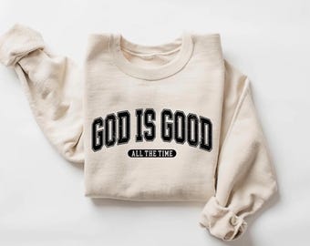 May include: A cream-colored sweatshirt with the text "GOD IS GOOD" in a black, collegiate-style font. Below the main text is the phrase "ALL THE TIME" in a smaller, black font. The sweatshirt is folded, with the sleeves visible.