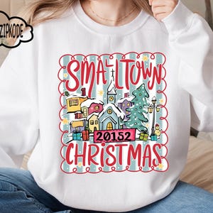 May include: White sweatshirt with a festive graphic. The design includes the words "SMALLTOWN CHRISTMAS" and the number "20152", alongside illustrations of a church, houses, a Christmas tree, and presents, all within a decorative frame.
