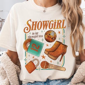 May include: Cream-colored t-shirt with a graphic that reads "Showgirl In My Showgirl Era." The design includes a disco ball, high-heeled boots, a record, a microphone, and a lock, all in glittery gold and orange.