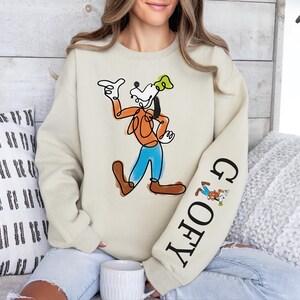 A woman wearing a white sweatshirt with a graphic of Goofy, a popular Disney character, sitting on a couch.