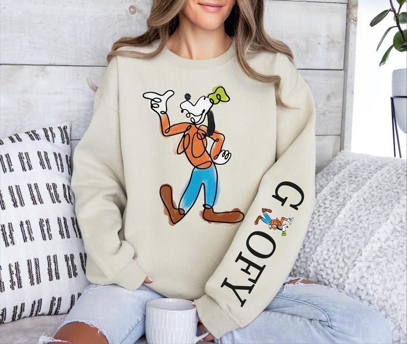 A woman wearing a white sweatshirt with a graphic of Goofy, a popular Disney character, sitting on a couch.