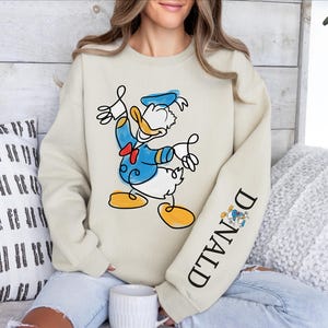 A woman wearing a sweatshirt with a cartoon image of Donald Duck on it, sitting on a couch with a coffee cup in front of her.