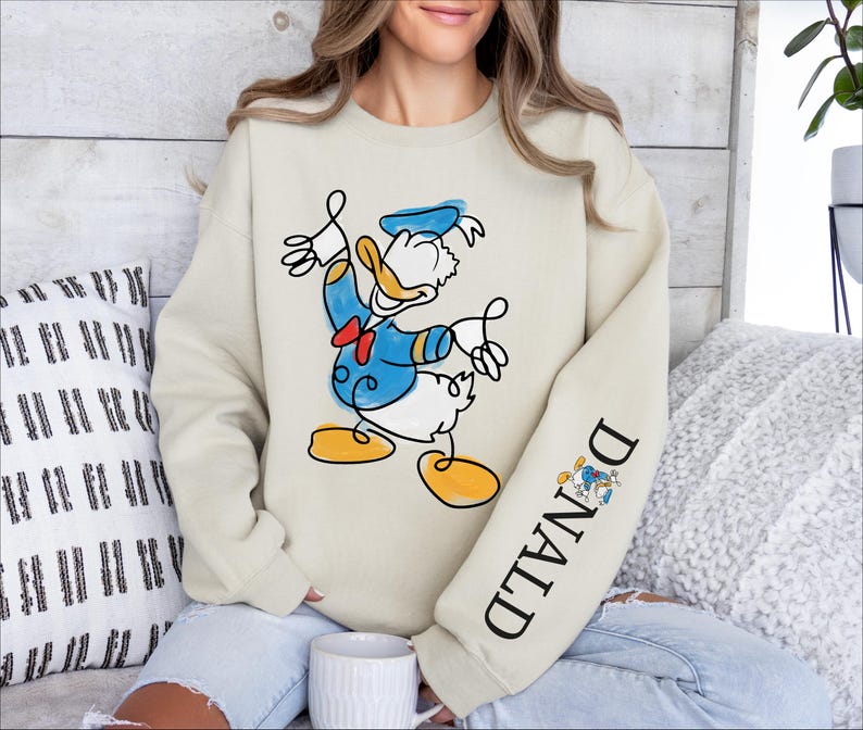 A woman wearing a sweatshirt with a cartoon image of Donald Duck on it, sitting on a couch with a coffee cup in front of her.