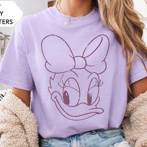 May include: A light purple t-shirt featuring a distressed graphic of Daisy Duck's face. The shirt has short sleeves and a crew neck. Text in the upper left corner reads "200+ DISNEY CHARACTERS".