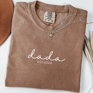 May include: A brown t-shirt with the text "dada est. 2024" in white lettering. The shirt is made of a soft, comfortable material.