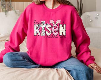 May include: A pink sweatshirt with a graphic design that says "He is Risen" with a cross and a bunny. The text "Matthew 28:6" is below the graphic.