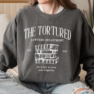 May include: Charcoal grey sweatshirt featuring the white text "THE TORTURED LAWYERS DEPARTMENT", a stack of books, and the phrase "All's fair in love and litigation". The sweatshirt has a relaxed fit.