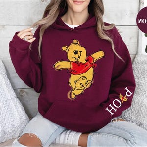 A woman wearing a red hoodie with a cartoon image of Winnie the Pooh on it, sitting on a couch.