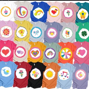 May include: A collection of colorful t-shirts featuring various cartoon character designs. Each shirt has a different graphic in a circular frame, including hearts, rainbows, and stars. The shirts come in a variety of colors, such as pink, blue, and yellow.