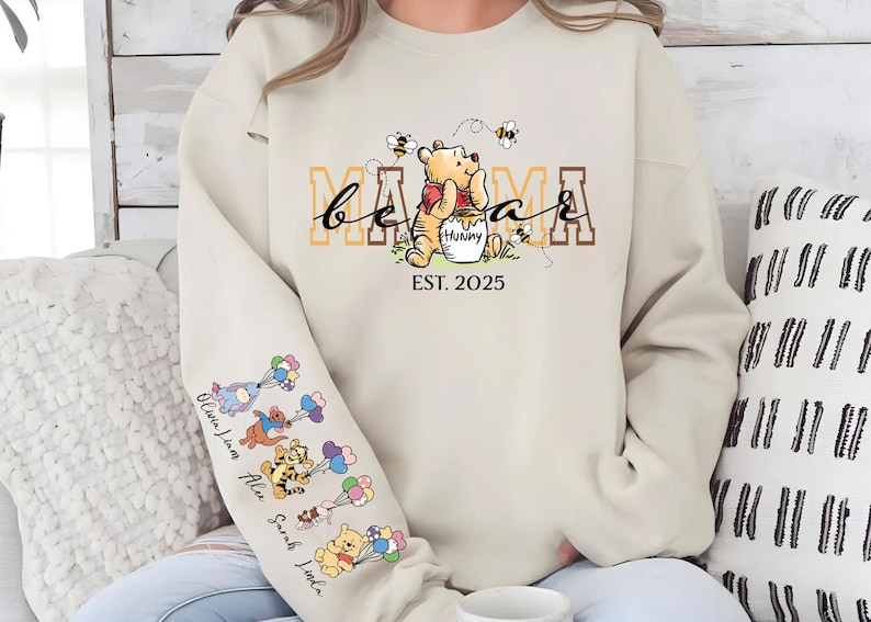A person wearing a white sweatshirt with a Winnie the Pooh-themed design, including the character's face and the text "Mama" and "Est. 2025".