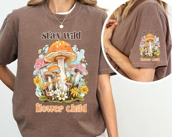 May include: Brown t-shirt with a graphic of mushrooms and flowers. The text "stay wild flower child" is printed on the shirt.
