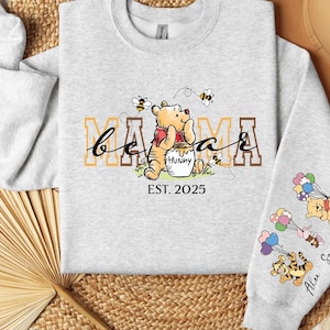 A gray sweatshirt with a Winnie the Pooh-themed design, including the character's face and the text "Mama" and "Est. 2025". The sweatshirt is laid out on a woven surface, with a hat and a fan visible in the background.