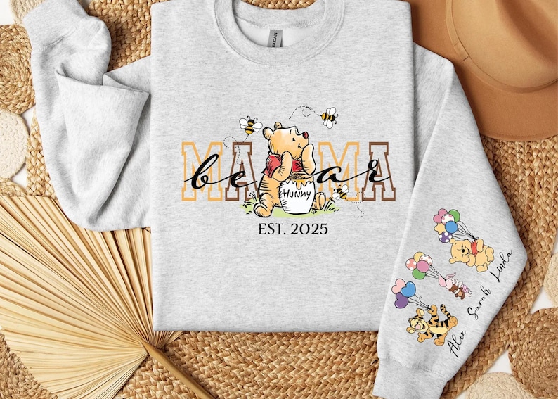 A gray sweatshirt with a Winnie the Pooh-themed design, including the character's face and the text "Mama" and "Est. 2025". The sweatshirt is laid out on a woven surface, with a hat and a fan visible in the background.