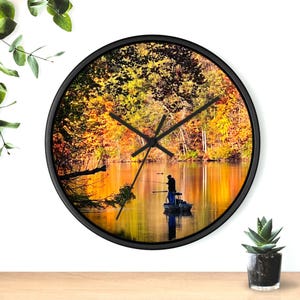 May include: A black round clock featuring a picture of a lake surrounded by autumn foliage. Two people are fishing from a boat in the lake. The clock hands are set to 10:10.
