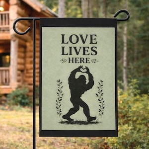 May include: A black garden flag featuring the text "LOVE LIVES HERE" and a silhouette of Bigfoot making a heart shape with its hands. The flag has a pale green background and is framed in black. Decorative plants flank the Bigfoot image.